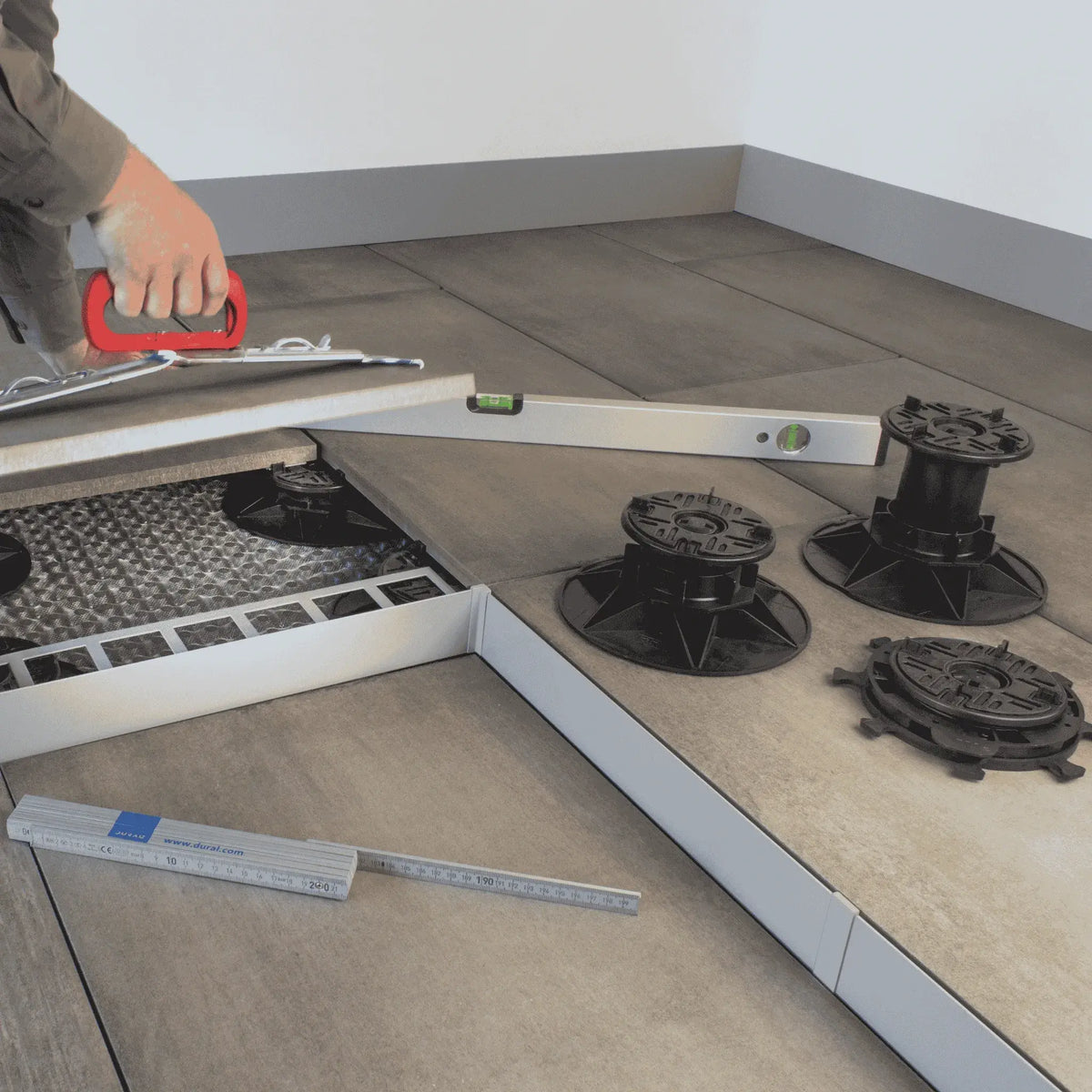 Ambientebild der 3 verschiedenen Stelzlagergrößen auf einer Terrasse. Der Handwerker benutzt eine Wasserwaage und einen Plattenheber zum Fliesenlegen. #A0005386 #A0005387 #A0005388